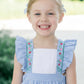 Young girl wearing a blue checkered dress with ruffled sleeves and floral embroidery, standing outdoors.