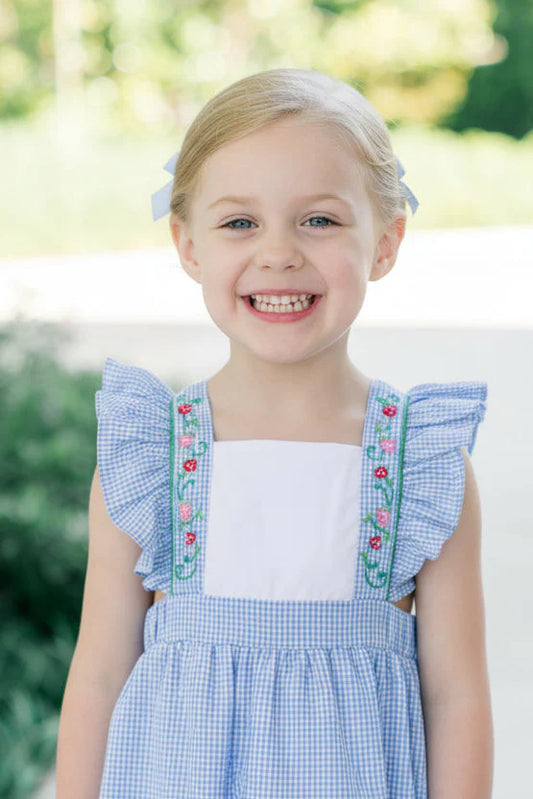 Young girl wearing a blue checkered dress with ruffled sleeves and floral embroidery, standing outdoors.