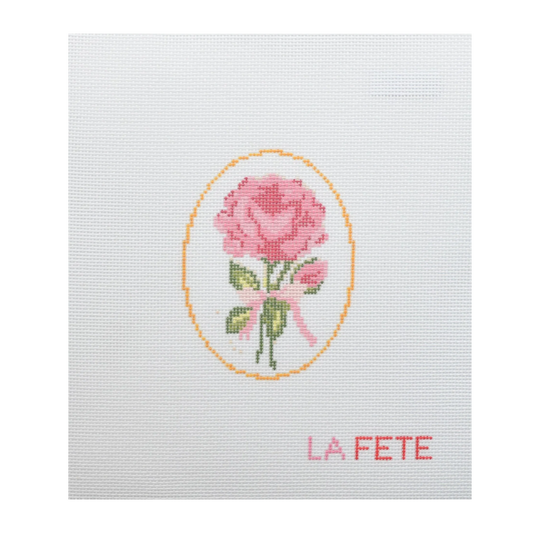 Cross-stitched design of a pink rose with a yellow border on white fabric, featuring the word 'LA FETE'.