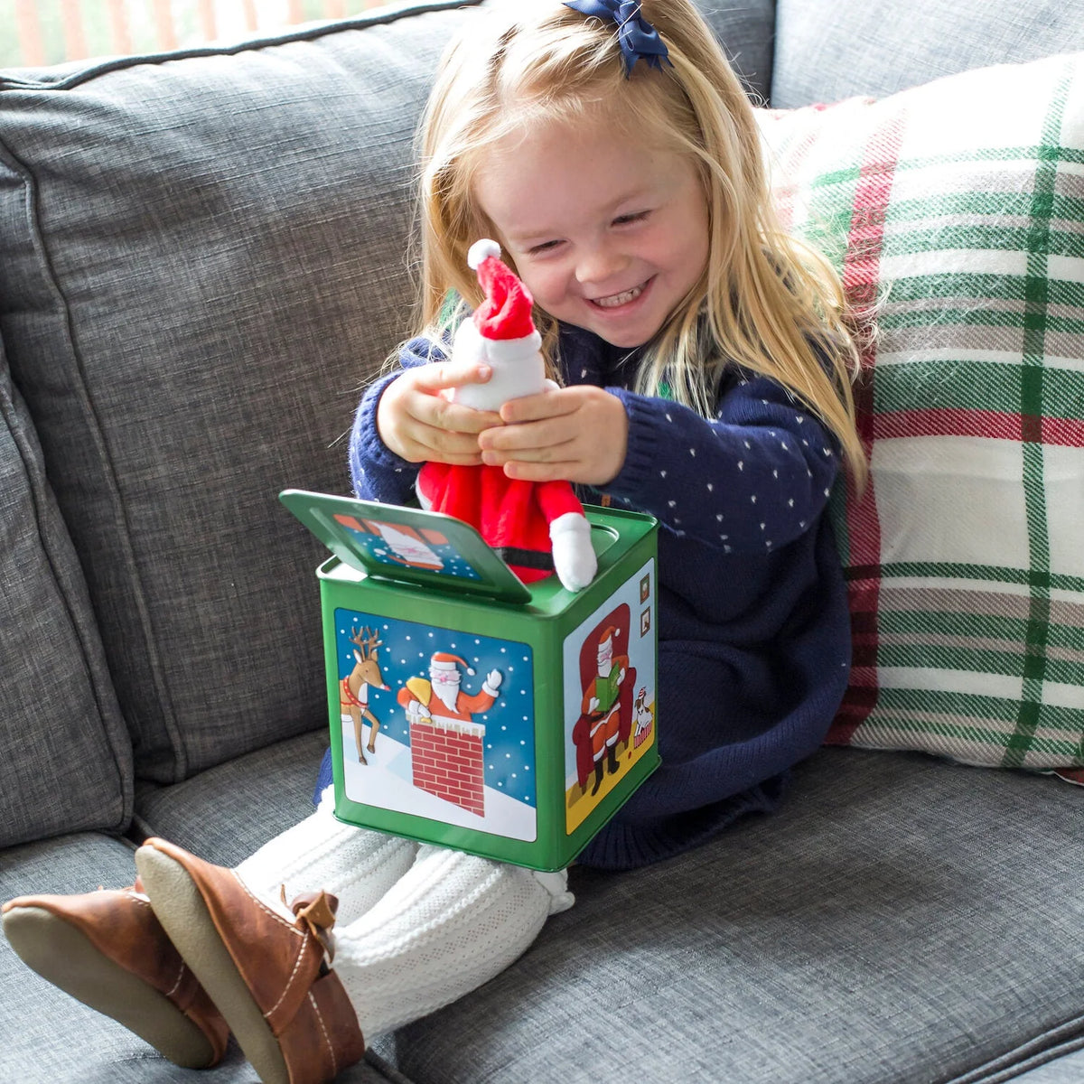 A young girl with blonde hair and a blue bow sits on a gray couch, smiling while playing with a Christmas-themed jack-in-the-box toy. The green box has colorful illustrations of Santa Claus and a train, and a Santa figure pops out of the top.