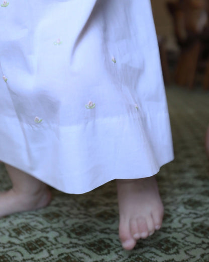 Close-up of a child's feet wearing a white dress with floral buttons on a patterned carpet.