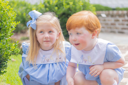 Two children in matching blue dresses with embroidered names, sitting outdoors.