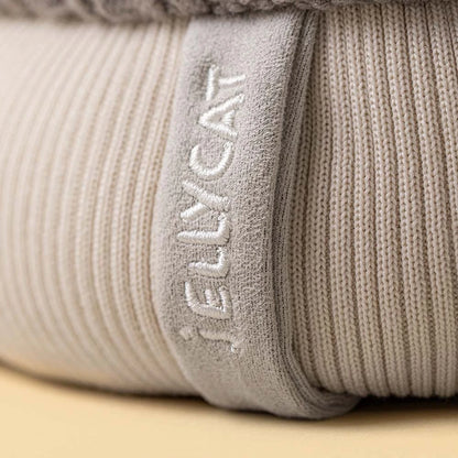 Close-up of a ribbed knit blanket or cushion in a light warm gray-beige color, with the brand name 'JELLYCAT' embroidered vertically on a fabric tab.