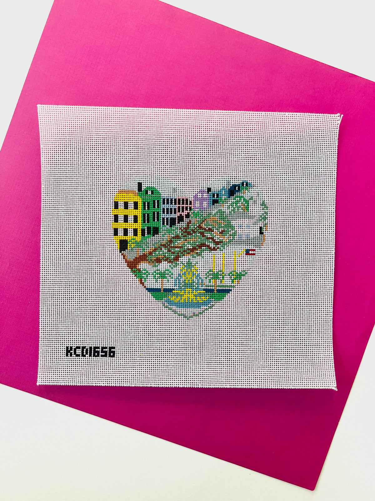 A square white cross-stitch canvas featuring a heart-shaped stitched cityscape with colorful buildings and greenery, placed on a bright pink sheet of paper. The black text 'KCD1656' appears in the lower left corner of the canvas.