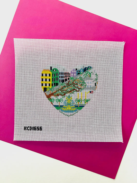 A square white cross-stitch canvas featuring a heart-shaped stitched cityscape with colorful buildings and greenery, placed on a bright pink sheet of paper. The black text 'KCD1656' appears in the lower left corner of the canvas.