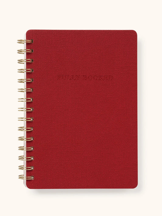 A red spiral-bound notebook with a textured cover and gold-toned wire binding, shown on a light cream background.