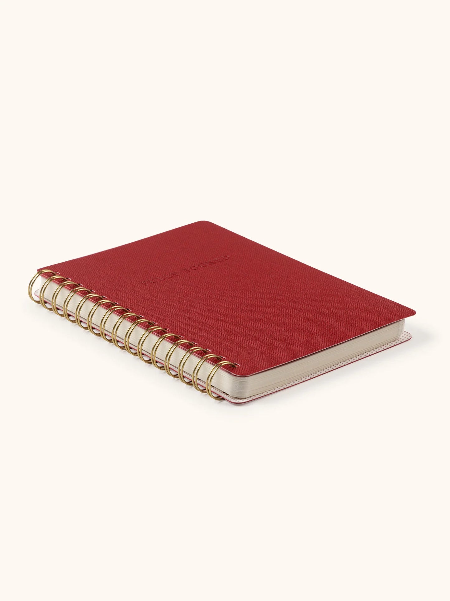 A small red spiral-bound notebook with a gold-toned wire binding and a light beige cover, photographed on a clean off-white background.