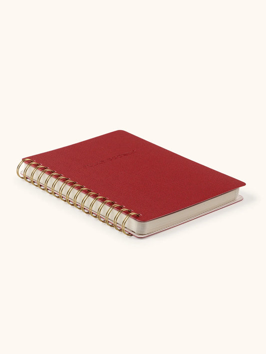A small red spiral-bound notebook with a gold-toned wire binding and a light beige cover, photographed on a clean off-white background.
