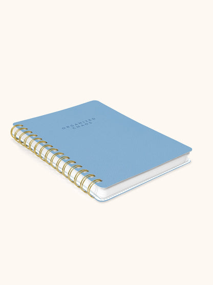A light blue spiral-bound notebook with gold wire binding and the words 'ORCHIDED STARS' printed on the cover, shown on a warm off-white background.
