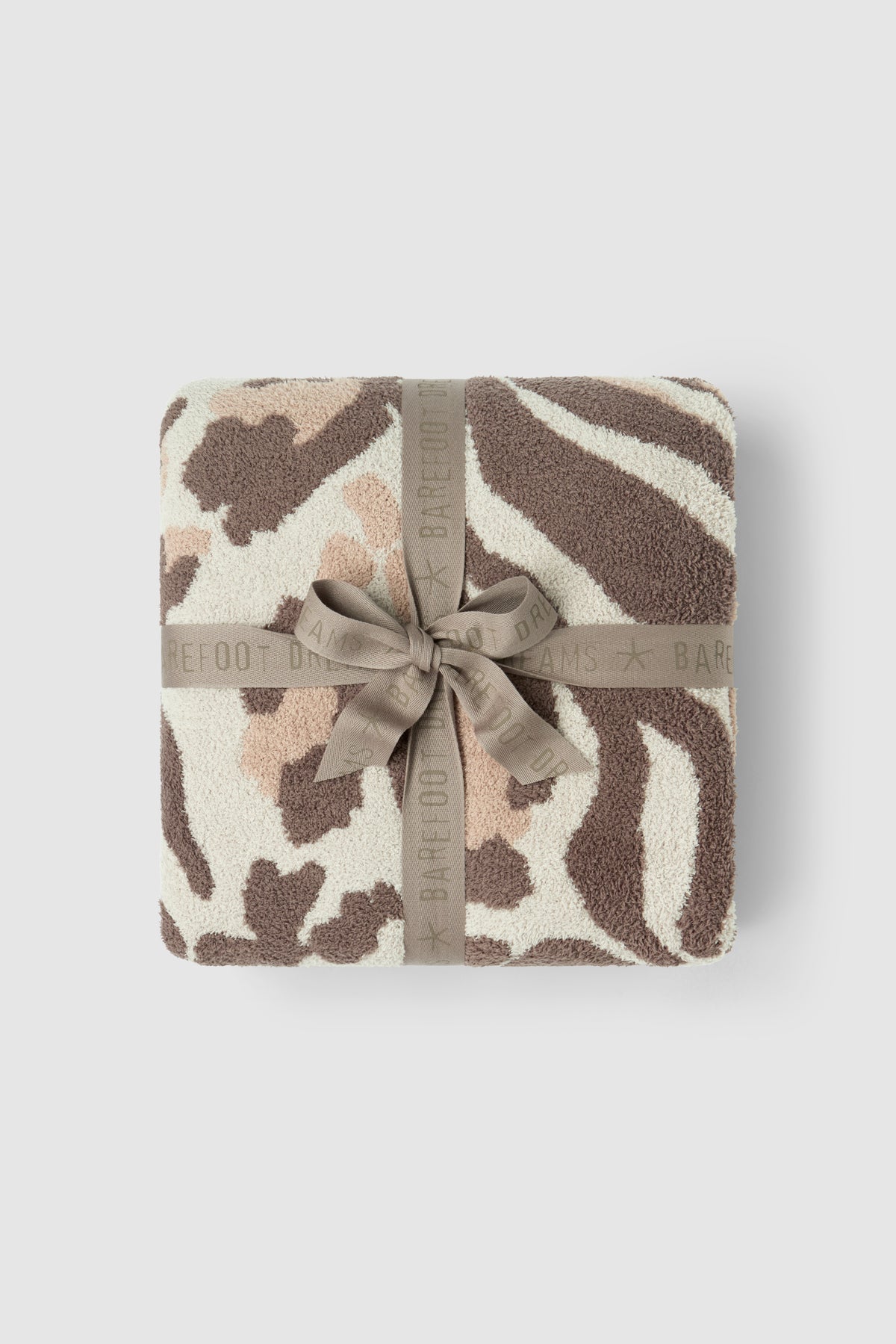 A folded plush throw blanket with a zebra-like animal print pattern in brown, beige, and cream, wrapped with a taupe ribbon that has the BARFOOT BAR brand name printed on it, set against a light gray background.