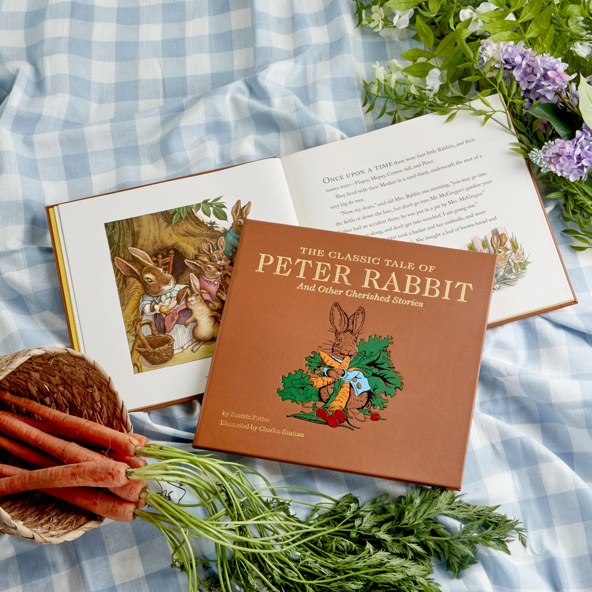 A styled flat-lay of the children's book 'The Classic Tale of Peter Rabbit' on a blue-and-white checkered blanket, with an open copy of the book behind it, carrots in a basket, and flowers around it.