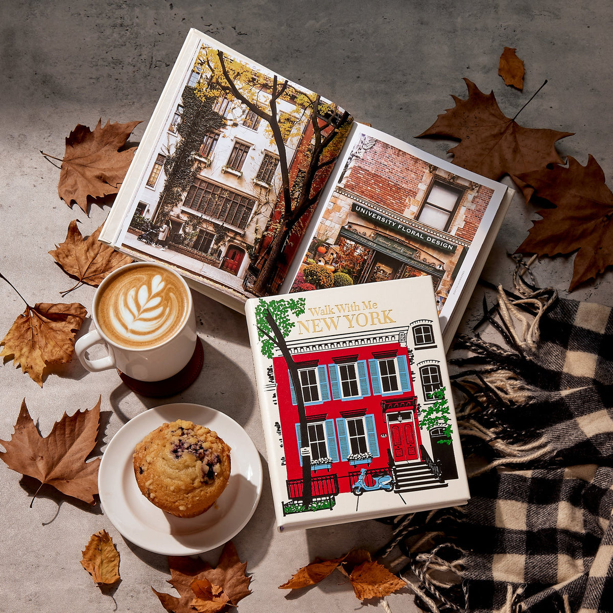 A styled flat-lay featuring a hardcover book titled 'You With Me, New York' in the foreground, with an open matching illustrated book behind it. The scene includes a cup of latte with leaf art, a muffin on a saucer, scattered autumn leaves, and a black-and-white plaid scarf on a gray surface.