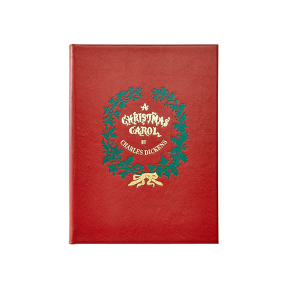 A red hardcover book featuring a green holly wreath and gold foil lettering that reads 'A Christmas Carol by Charles Dickens' on the front cover, shown against a plain white background.