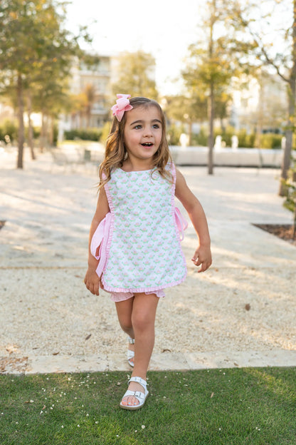 A young girl wearing a sleeveless dress with a turtle print and ruffle trim, in pink and white colors, walking on grass.