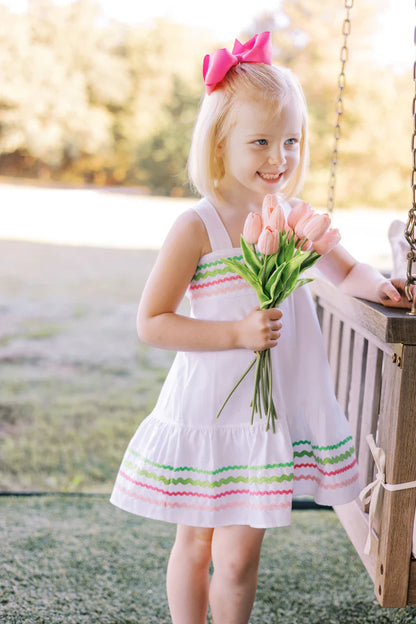A white Sundress with multicolored ric rac trim and a bow at the back neck, designed for children.