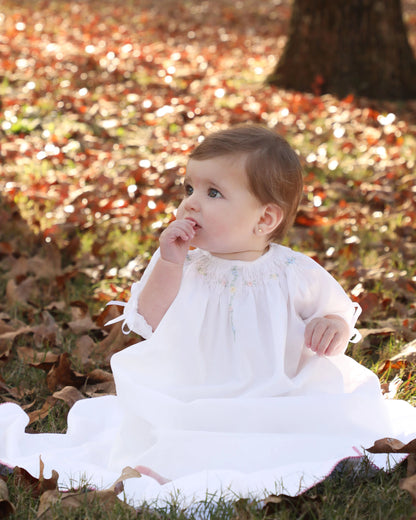 Baby in a white dress sitting on the ground with autumn leaves in the background