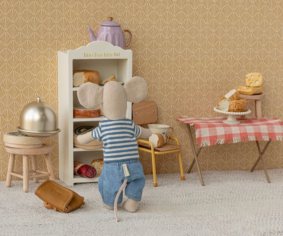 Miniature dollhouse scene with a child-like figure, shelves, and table with food items.