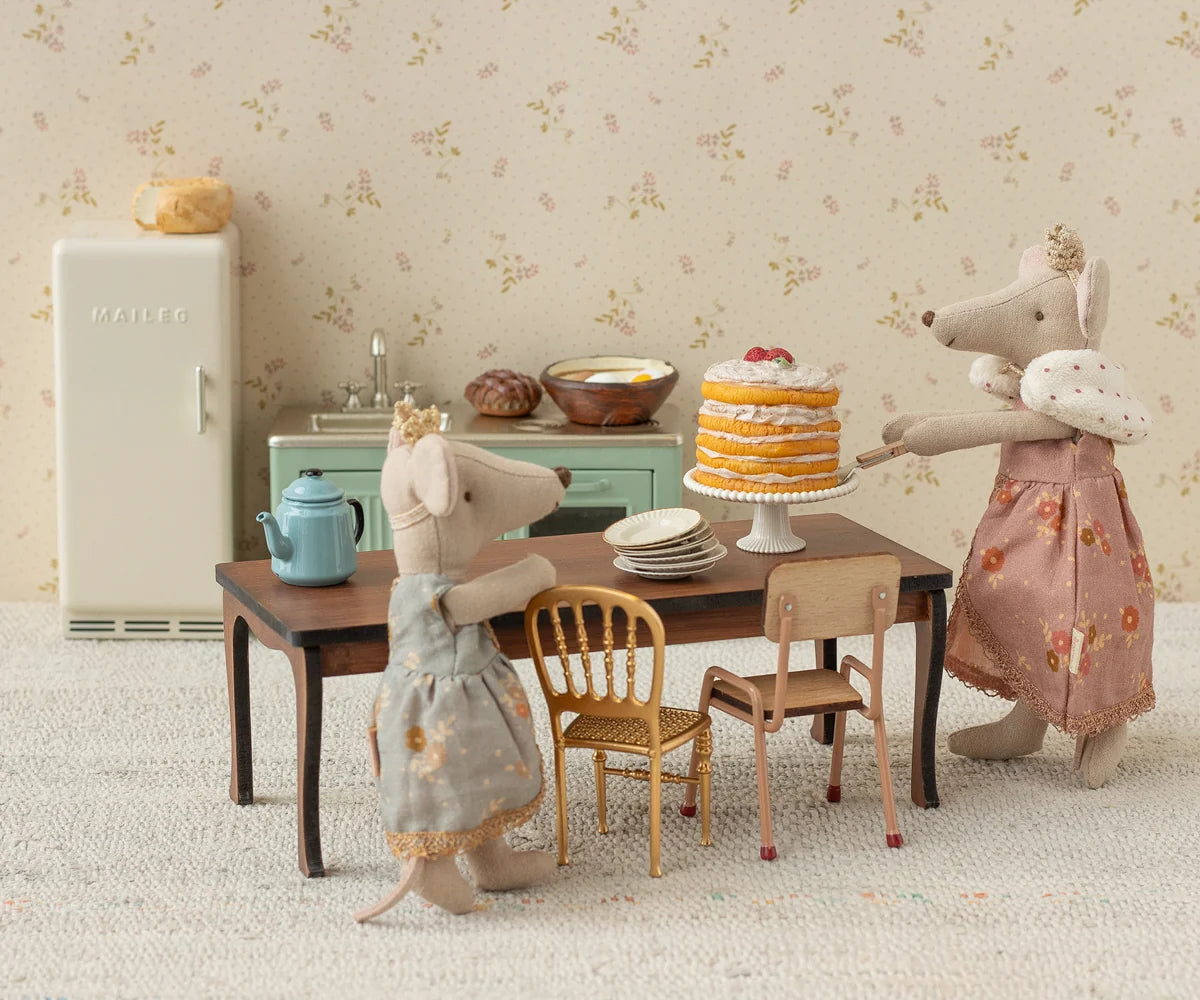 Two toy mice in dresses standing at a table with a cake and tea set, in front of a floral wallpapered wall.