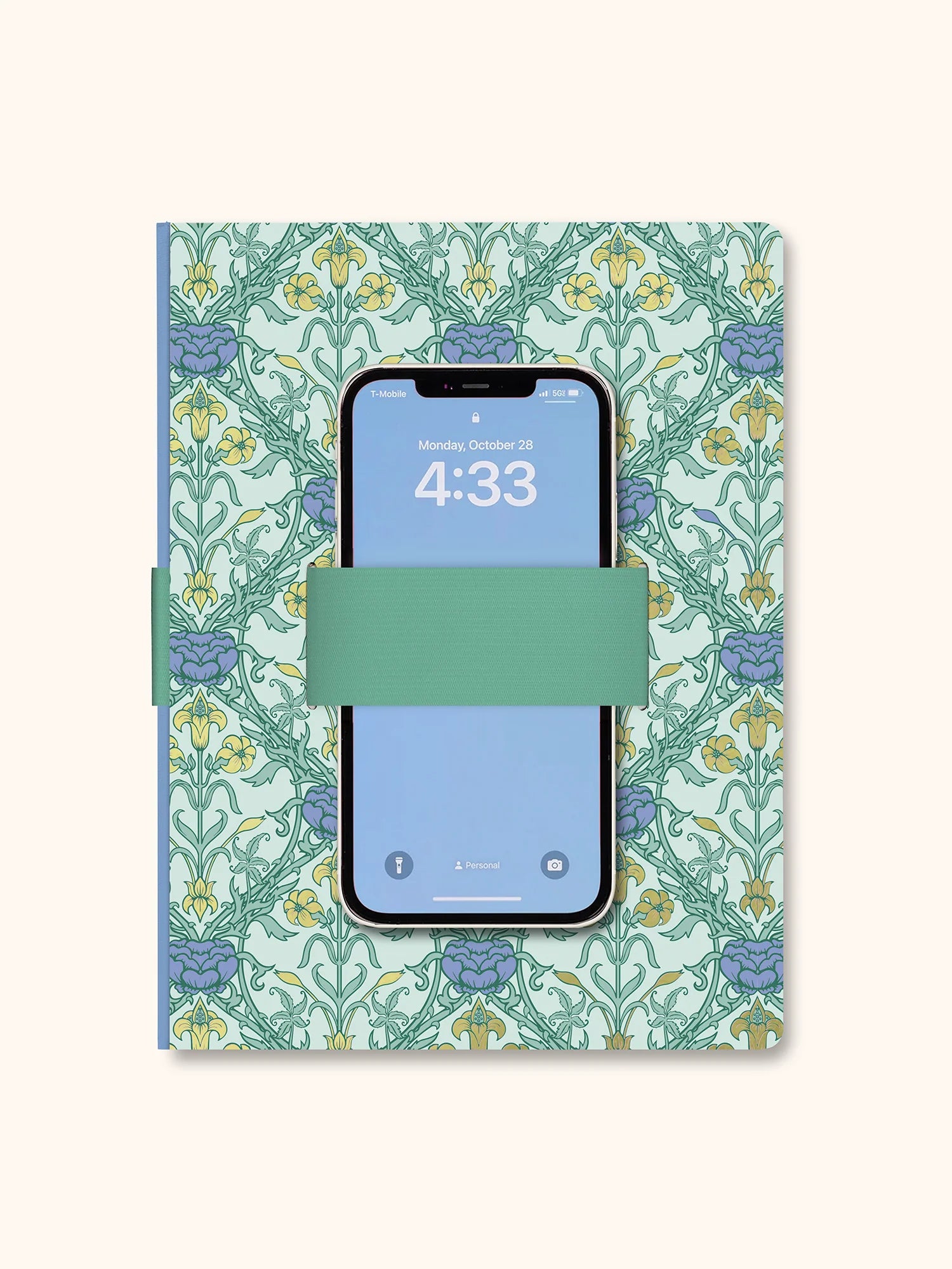 A notebook with a light green and blue floral patterned cover, featuring a mint green elastic band across the middle holding a smartphone. The notebook is photographed flat against a solid cream background.