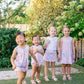 Four young girls in matching dresses standing on grass with trees in the background