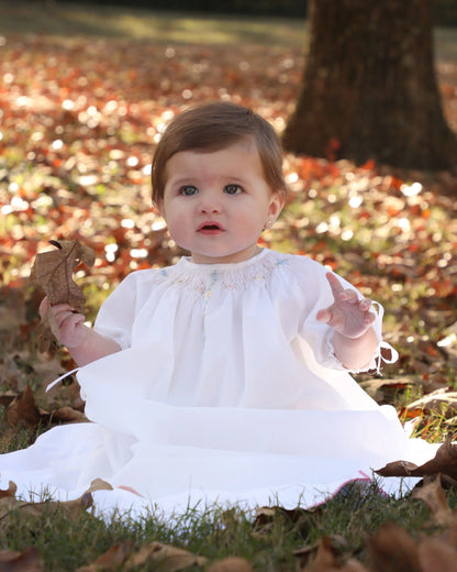 Baby in a white dress sitting on the ground with autumn leaves in the background