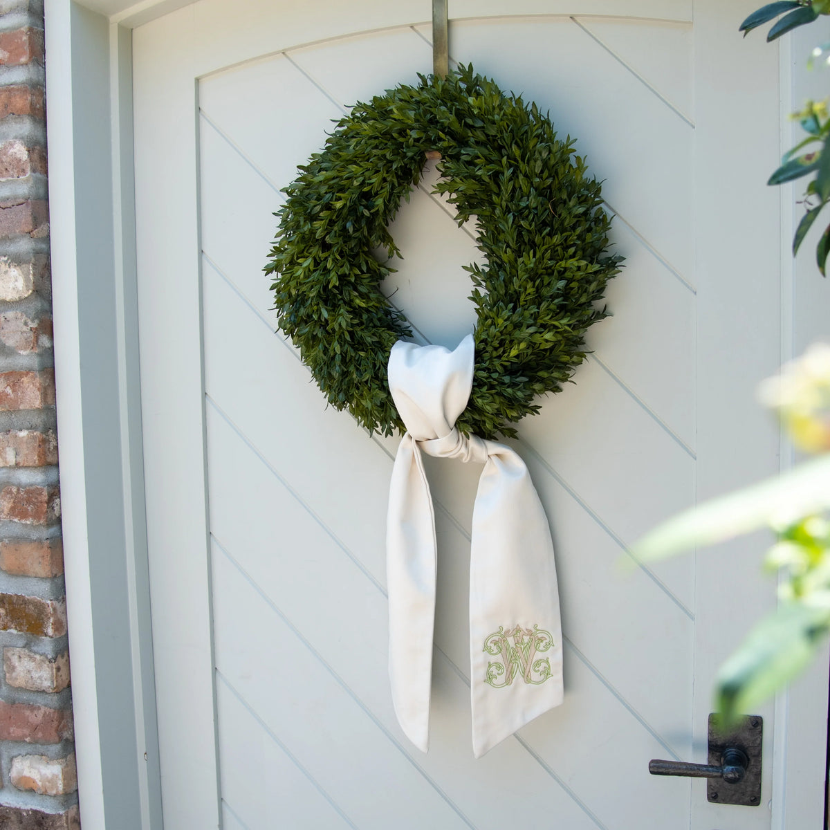 A green boxwood wreath hanging on a white paneled front door, tied with a white satin bow featuring a gold monogram.