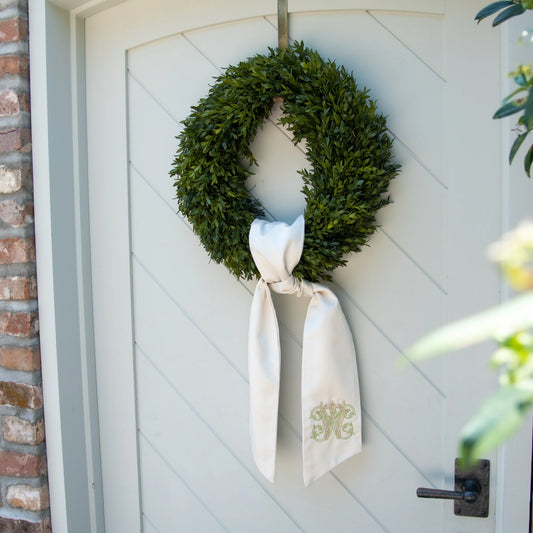 A green boxwood wreath hanging on a white paneled front door, tied with a white satin bow featuring a gold monogram.