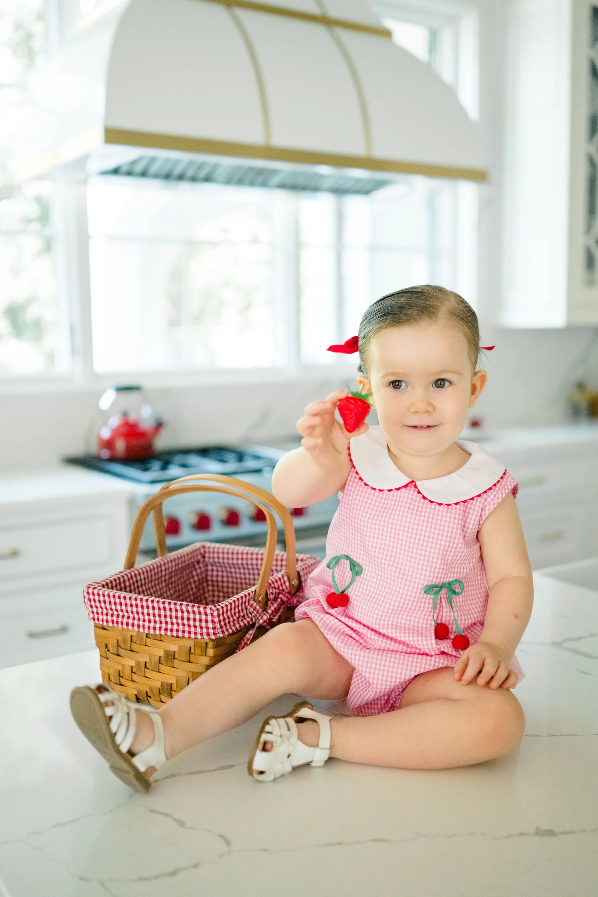 Baby in a pink outfit with red cherries holding a cherry in a kitchen.