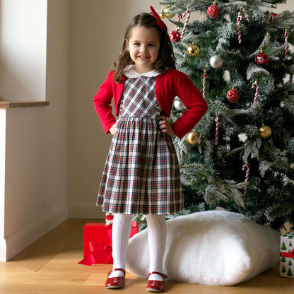 Young girl in a plaid dress and red cardigan standing in front of a decorated Christmas tree.