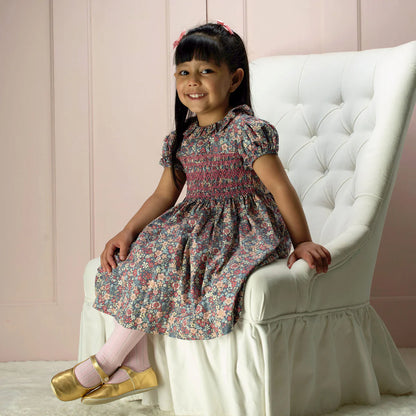 Young girl in a floral dress sitting on a white chair against a pink wall.