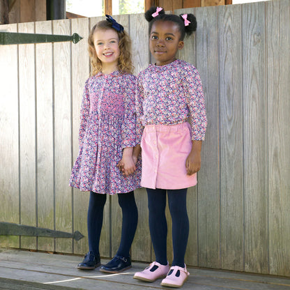 Two young girls in matching floral tops and pink skirts standing in front of a wooden fence.