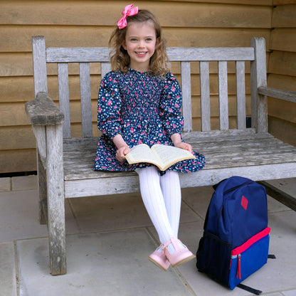 Young girl sitting on a wooden bench with a book, wearing a floral dress and pink shoes, with a blue and red backpack next to her.