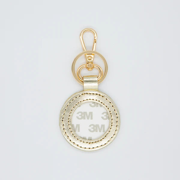 A gold-colored metal keychain accessory featuring a carabiner clip, two interlocking rings, and a textured circular ring with a clear protective cover showing 3M adhesive backing.