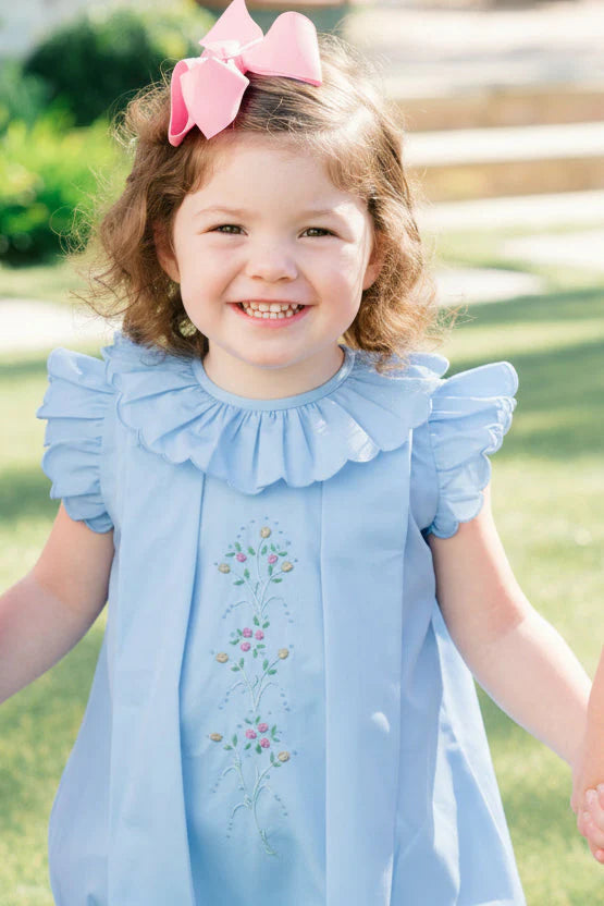 Young girl in a light blue dress with a pink bow in her hair, standing outdoors.