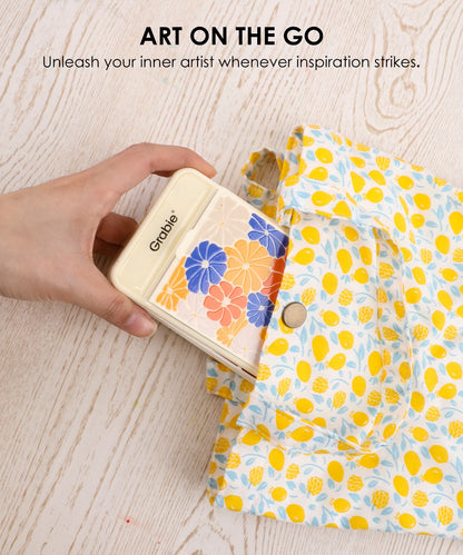 Hand holding a small wooden art kit with floral designs, next to a colorful fabric pouch on a light wood surface.