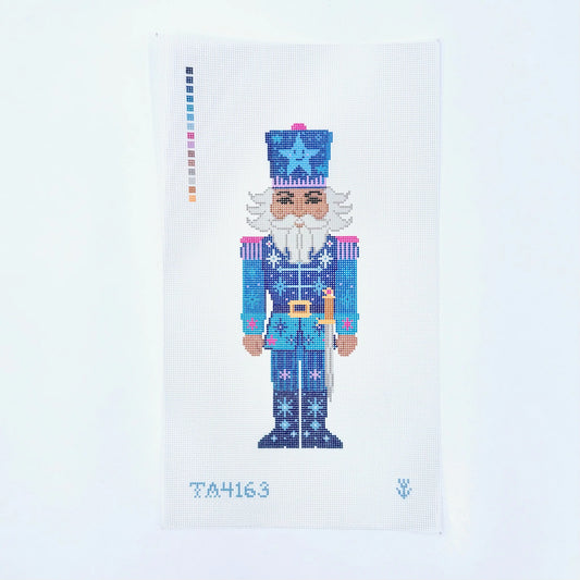 A needlepoint canvas featuring a pixelated nutcracker design in shades of blue, white, and pink, with a color palette strip on the left and the number TA4163 at the bottom.