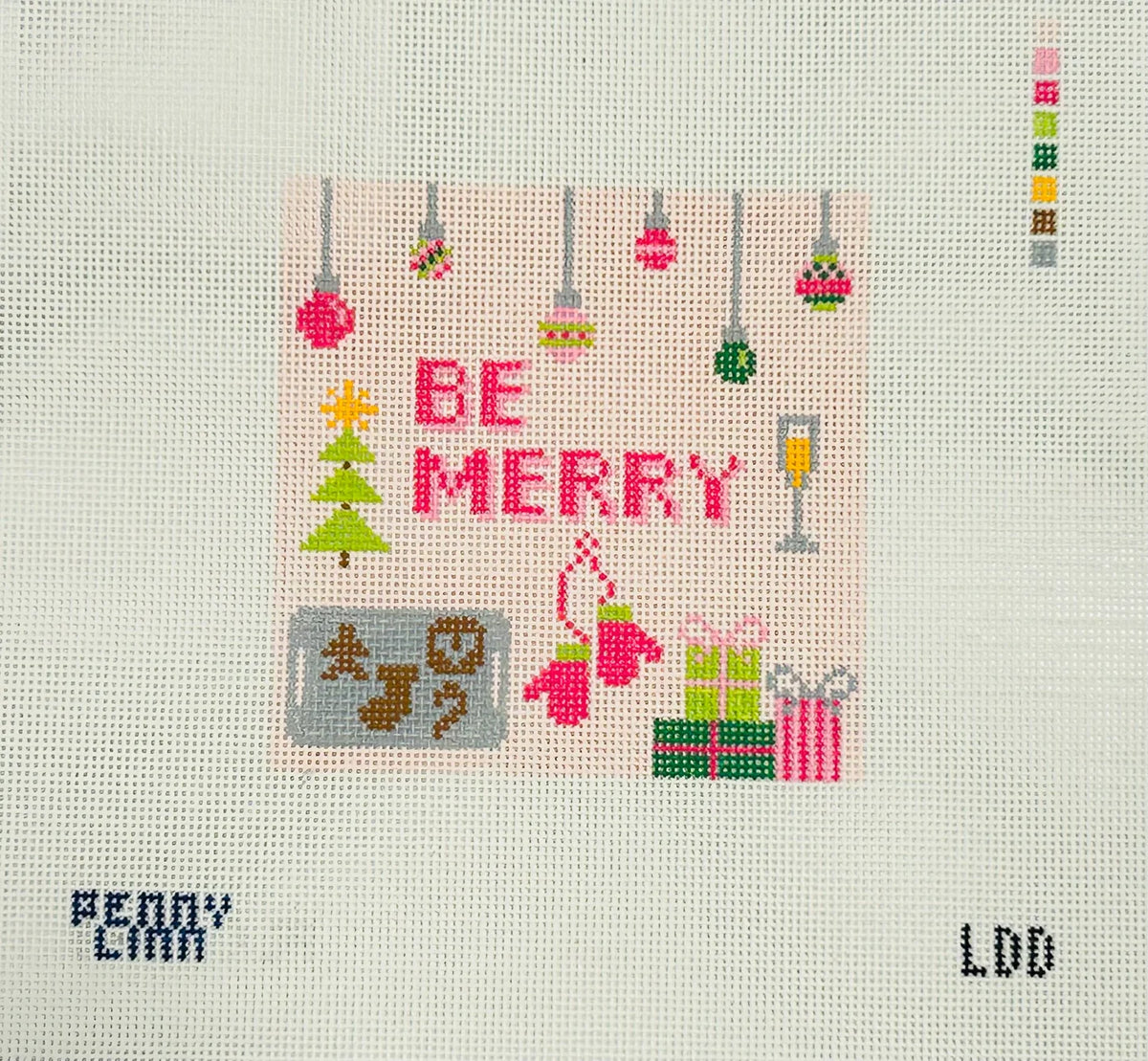 A square needlepoint canvas featuring a festive Christmas design with the words 'BE MERRY' in pink, a green tree, ornaments, gifts, and holiday motifs on a white mesh background.