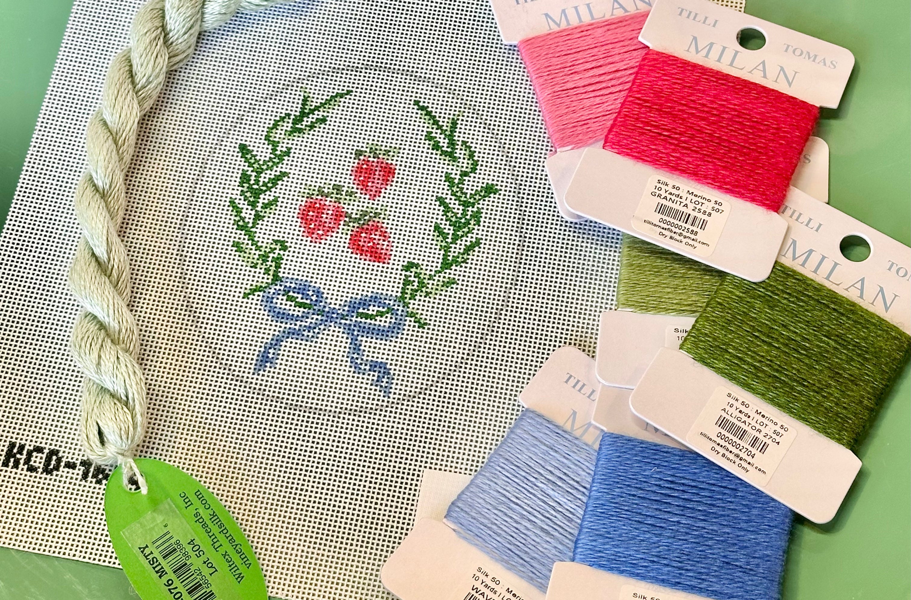 Needlepoint canvas with strawberry design and colorful thread packages on a green surface