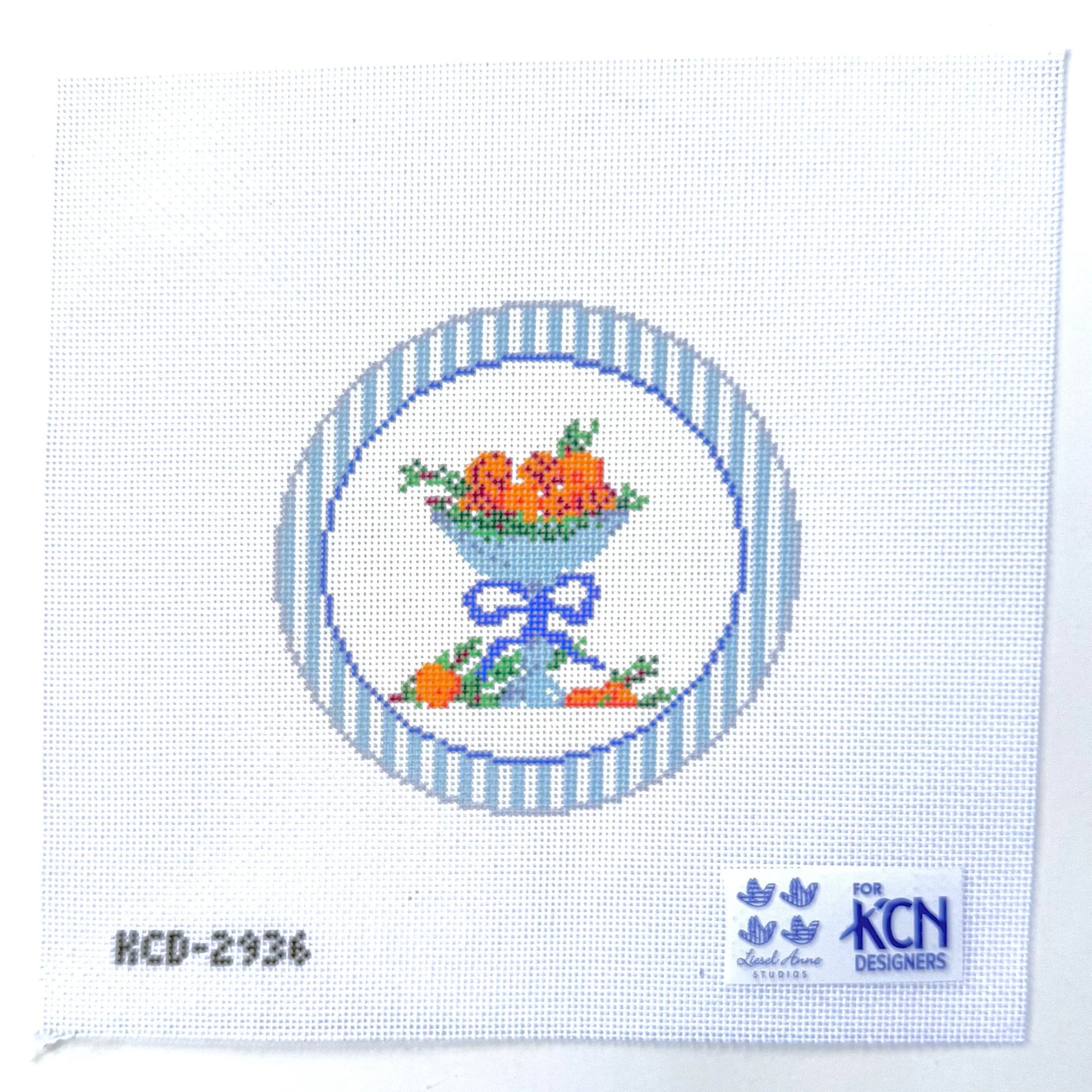 Cross-stitch pattern with a blue bowl and fruit design on white fabric, featuring KCN Designers logo.