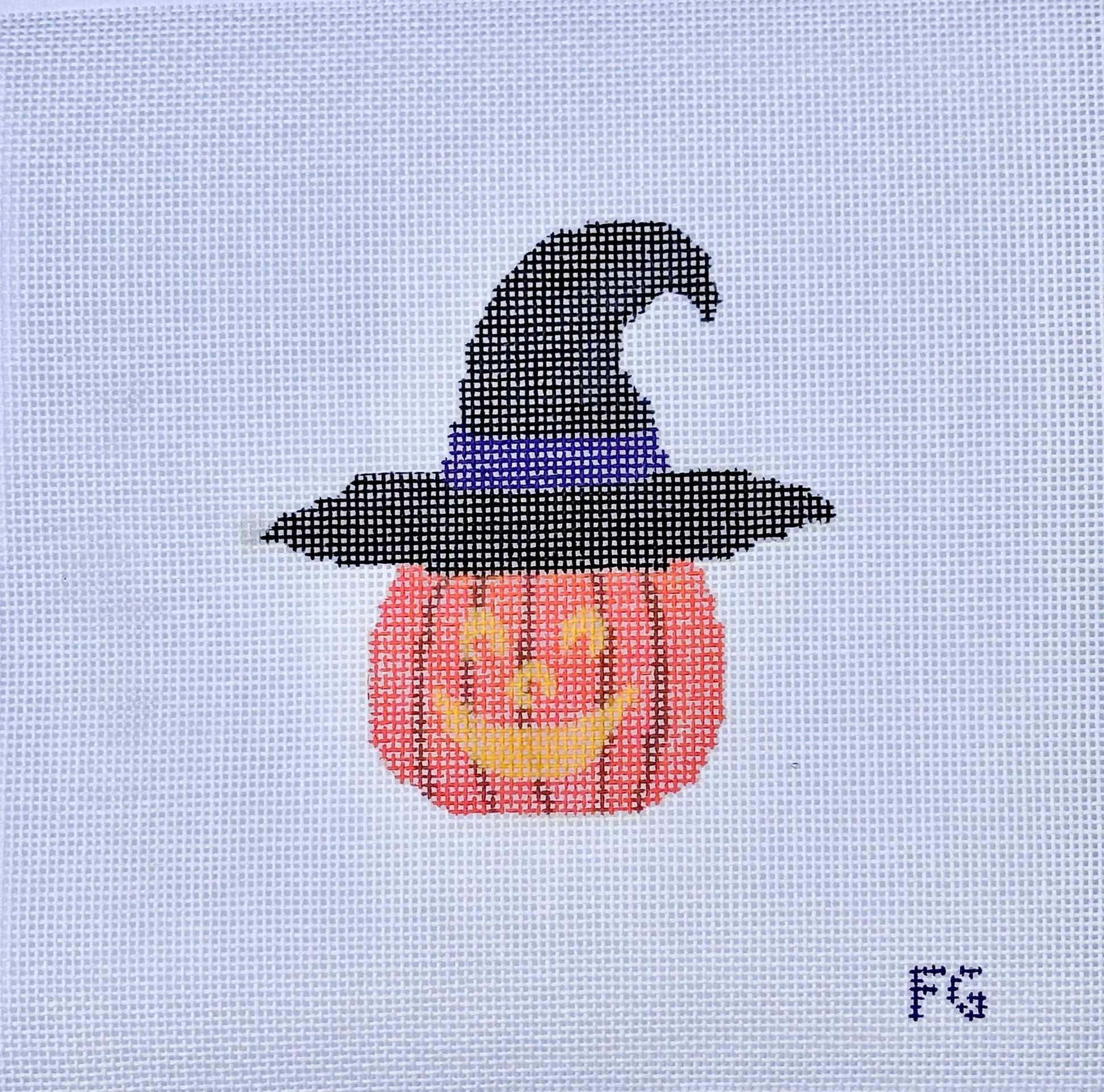 A needlepoint canvas featuring a pixelated design of a jack-o'-lantern pumpkin wearing a black witch hat with a purple band, on a white mesh grid. The initials 'FG' are printed in the bottom right corner.