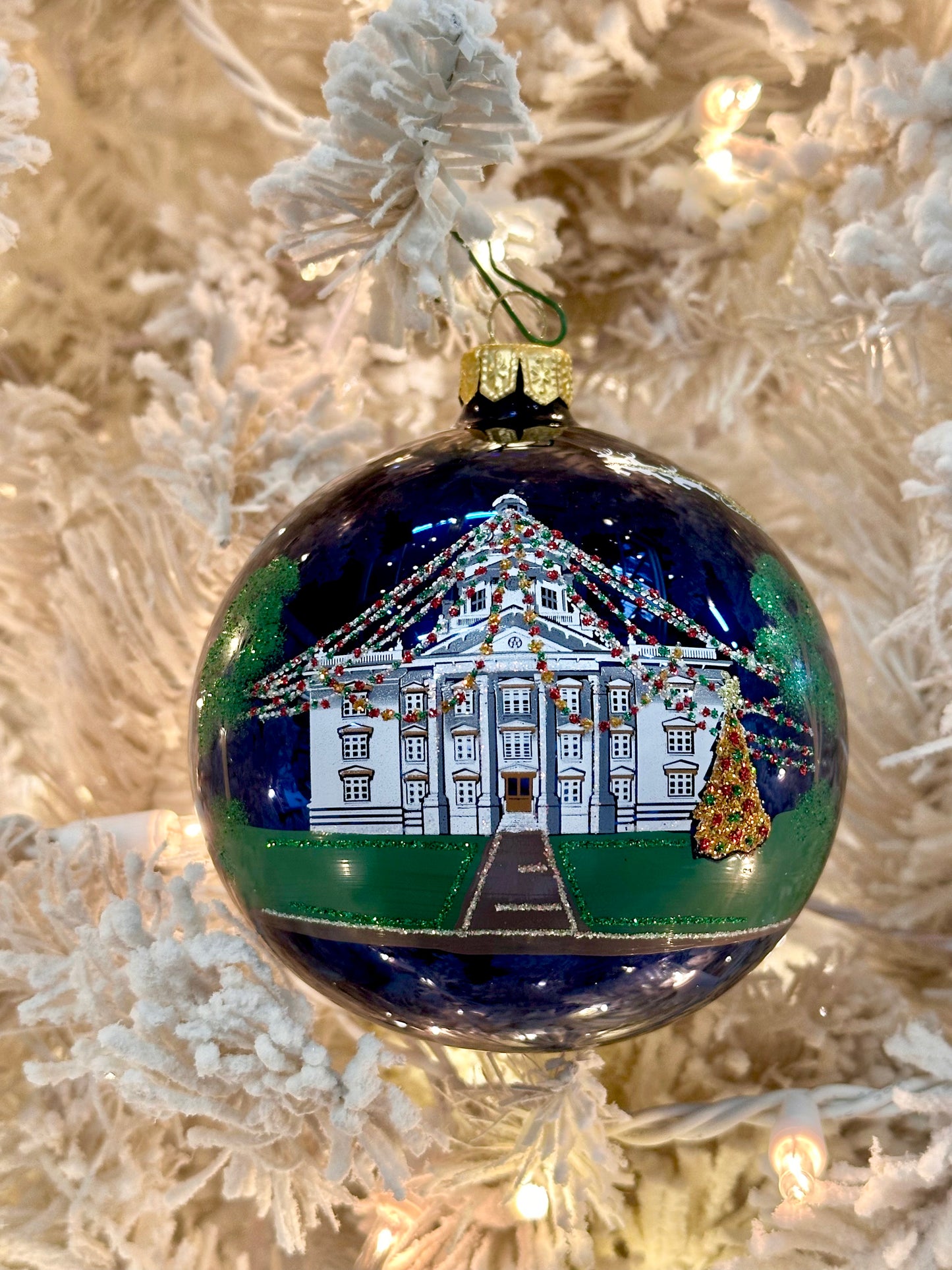 A round glass Christmas ornament featuring a hand-painted white courthouse decorated with colorful Christmas lights and a small gold Christmas tree, hanging on a white flocked Christmas tree with warm string lights.