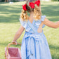 Young girl in a blue dress with red bows in her hair, holding a picnic basket outdoors.