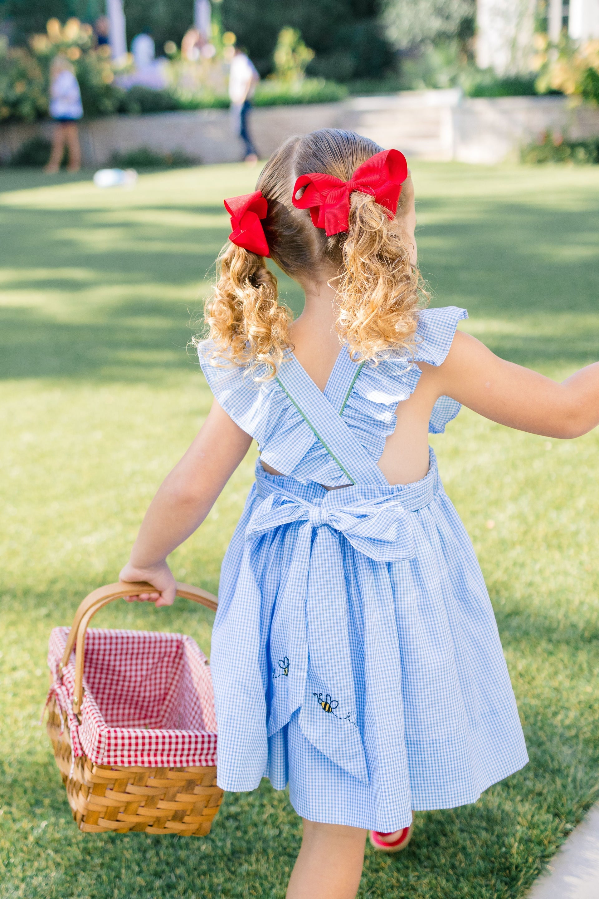 Young girl in a blue dress with red bows in her hair, holding a picnic basket outdoors.