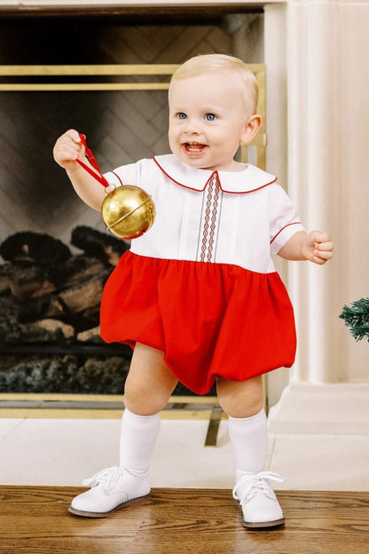 Baby in a red and white dress holding a gold bell indoors.
