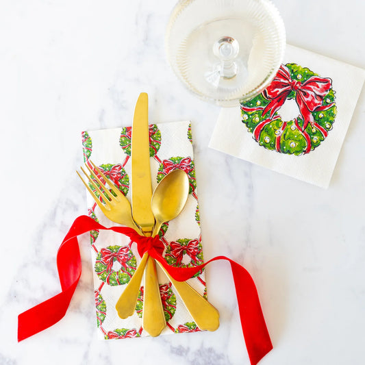 A styled holiday table setting on a white marble surface, featuring a white napkin printed with green wreaths and red bows, tied with a red ribbon and topped with gold flatware. A matching printed napkin and a clear glass plate are arranged nearby.