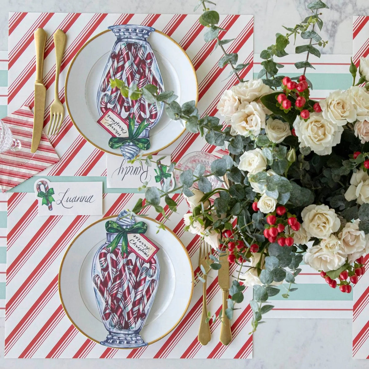 A styled holiday table setting featuring two white plates with gold trim, printed with an illustration of a blue-and-white vase filled with red-and-white candy canes. The plates are set on red-and-white striped placemats with gold-toned flatware, place cards, and a large floral centerpiece with white roses, green eucalyptus, and red berries.