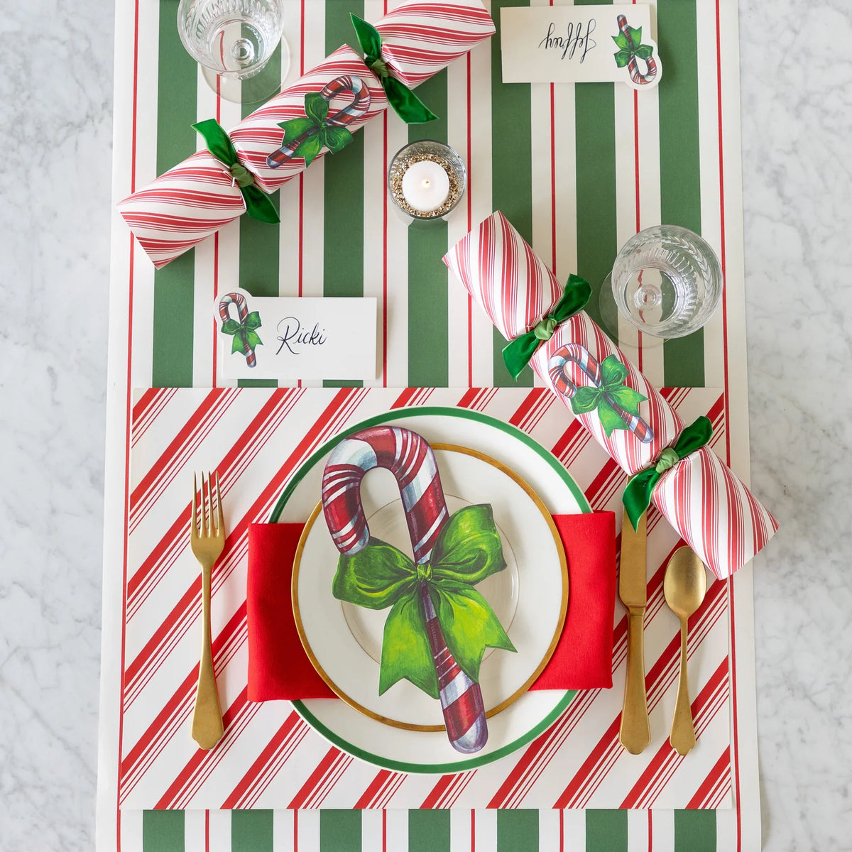 A festive holiday table setting featuring a green and white striped table runner, red and white striped napkins tied with green ribbon, a white plate with a candy cane and green bow print, gold cutlery, and place cards on a light marble surface.