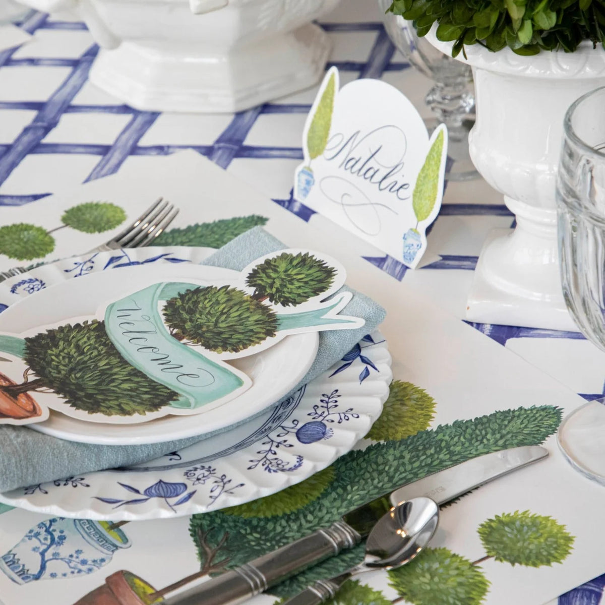 A styled tabletop place setting with white plates, blue floral patterns, silver cutlery, green textured napkins, and decorative paper toppers reading 'welcome' and 'Nature'.