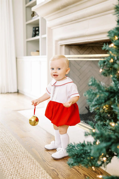 Baby in a red and white outfit standing near a Christmas tree and fireplace.