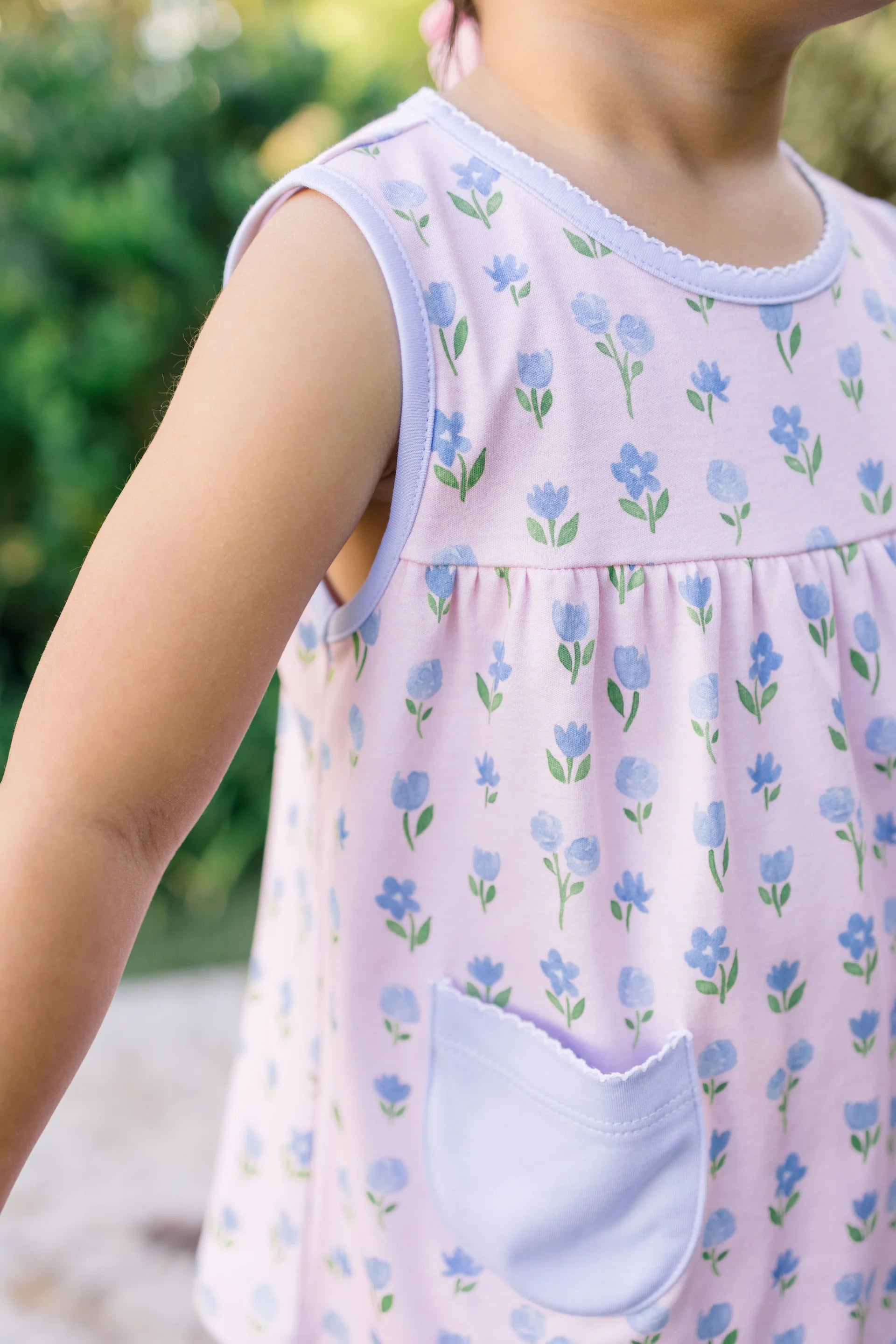 Child wearing a sleeveless dress with floral pattern and pocket, outdoors.